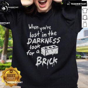 Official 'When You're Lost In The Darkness Look For A Brick' sweatshirt displayed on a hanger, showcasing its unique design.