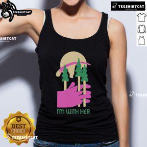 Alt Text: Woman wearing a 'Nice, I'm With Her' tank top surrounded by three trees in a sunny outdoor setting.