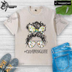 Mom enjoying a pretty camping life in a messy bun, wearing a washed T-shirt, surrounded by nature and camping gear.