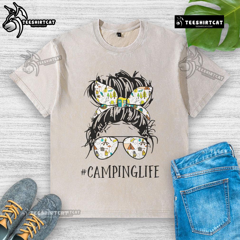 Mom enjoying a pretty camping life in a messy bun, wearing a washed T-shirt, surrounded by nature and camping gear.